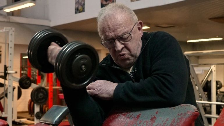 Jean-Pierre, lunettes et pull noir, appuyé sur un pupitre à biceps rouge, soulève un haltère dans la salle BodyGym